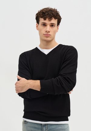 Giovane uomo con capelli castani e ricci che indossa un maglione nero a V sopra una camicia bianca, in piedi con le braccia incrociate davanti a uno sfondo semplice.