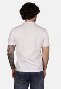 T-shirt bianco in cotone visto da dietro, con maniche corte e un design semplice, abbinato a jeans di jeans blu e tatuaggi visibili.