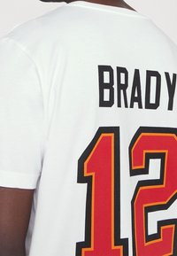 Bílý bavlněný tričko s černým textem "BRADY" a velkým červeným a žlutým číslem "12" na zádech. Krátké rukávy, kulatý výstřih.