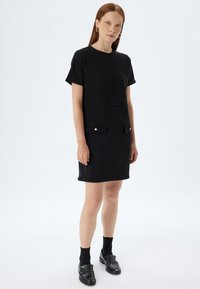 Zwarte jurk met korte mouwen, rechte vorm, voorzakdetails en accenten in goudkleurig metaal. Glad stofoppervlak.