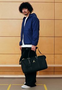 Saco desportivo preto com logótipo dourado, material liso, alças resistentes e detalhes de fecho éclair. Modelo veste um casaco acolchoado azul-marinho e calças pretas largas.