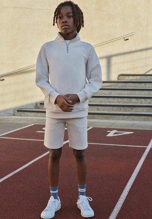 Lysebeige sweatshirt med lynlås og raglanærmer, kombineret med matchende shorts. Hvide sneakers og blåstribede sokker på en bane baggrund.