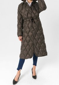 Lång, mörk olivgrön quiltad pufferjacka med huva, cinchad midja och tryckknappar fram; har diamants mönster och dragsko.