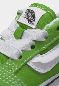 Nærbilde av tungen på en grønn Vans-sneaker med hvite lisser og "Vans Off The Wall"-logoetikett.