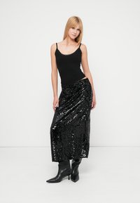 Jupe noire à sequins avec un design fluide, associée à un débardeur noir ajusté. Le mannequin porte des bottes noires à bout pointu.