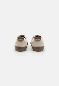 Sneaker Lacoste con tomaia beige, dettagli marroni e suola in gomma testurizzata. Presenta un logo sul tallone e un design minimalista.