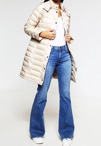 Zateplený béžový kabát s vysokým límcem a kopytovými knoflíky, kombinovaný s rozšířenými modrými džíny a bílými teniskami. Hladká textura, ležérní design.