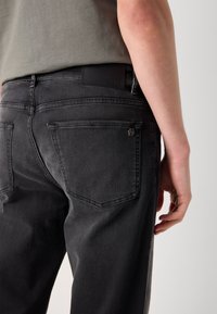 Personne portant un jean gris foncé et une chemise grise, montrée de la taille vers le bas avec un gros plan de la zone de la poche arrière.