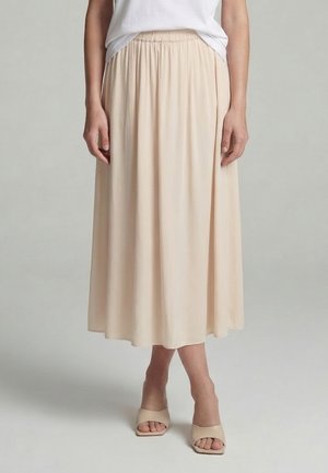 Woman wearing a beige midi skirt with an elastic waistband, white top, and beige open-toe heeled sandals, standing against a plain background.