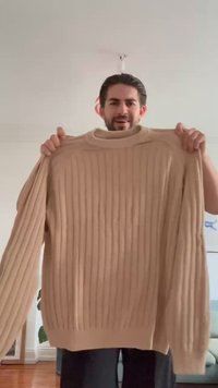 Beige gerippter Strickpullover mit hohem Kragen und langen Ärmeln, der ein strukturiertes Muster und eine lockere Passform aufweist.