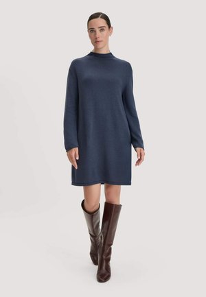 Vrouw die een marineblauwe sweaterjurk met lange mouwen en donkerbruine leren kniehoge laarzen draagt, staand op een lichte achtergrond.