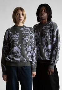 Dwie osoby ubrane w dopasowane szare swetry z dużymi abstrakcyjnymi wzorami czaszek, stojące obok siebie na jednolitym tle.