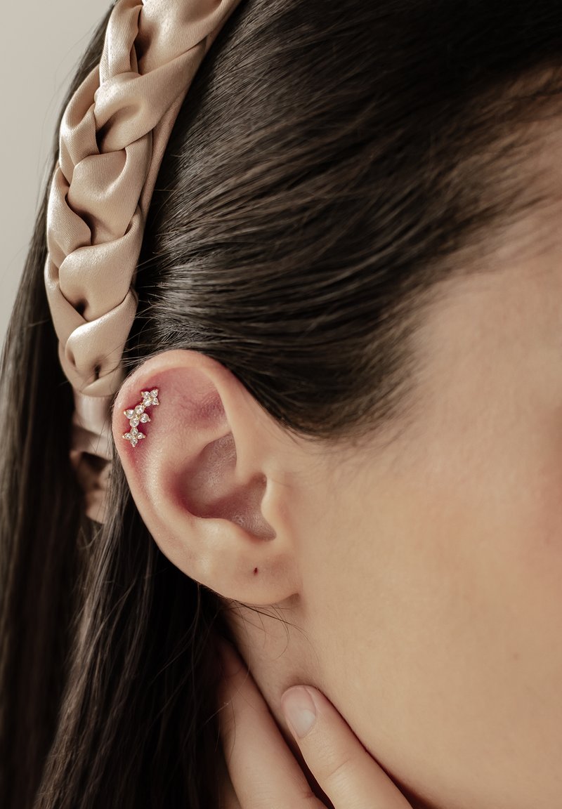 Joyería de piercing de oro y cristal con un diseño floral, sobre un tono de piel claro, resaltada por una diadema beige satinada.