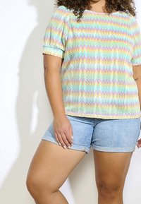 Top de manga corta con un patrón de zigzag multicolor, hecho de una tela ligera y texturizada. Se lleva con pantalones cortos de mezclilla, mostrando los dobladillos doblados.