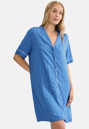 Kobieta w niebieskiej koszuli nocnej w paski do kolan z krótkim rękawem, zapinanej na guziki, z białą lamówką na kołnierzu i rękawach, stojąca na białym tle.