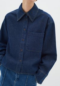 Dark blue denim shirt with a classic collar, button front, and left chest pocket. Features visible stitching and relaxed fit.