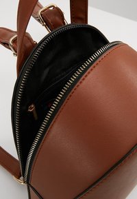 Pequeña mochila marrón de piel sintética con cierre de cremallera, forro interior negro y detalles en dorado. Incluye un bolsillo interior para almacenamiento.