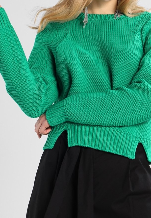 Femme portant un pull en maille verte texturée à manches longues et une jupe plissée noire sur un fond uni.