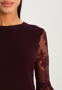 Bordeaux top met lange kanten mouwen met bloemenpatronen, geribbelde textuur op het lichaam en een ronde halslijn.