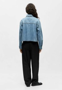 Denim jakna svijetlo plave boje s krojem koji se skraćuje, s dugmadima na manžetama i klasičnim okovratnikom, u kombinaciji s crnim širokim hlačama.