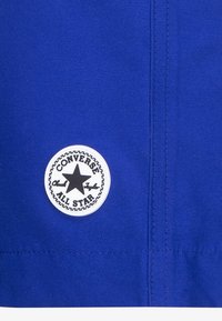 Blått stoff med en rund hvit lapp med en svart stjerne og teksten "CONVERSE ALL STAR" i en sydd kant. Glatt tekstur, uformelt design.