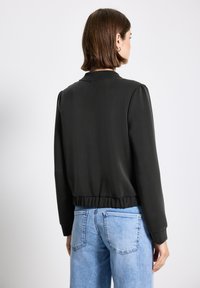 Musta bomber-takki, jossa on ribbireunus ja puffatut hihat, silkkinen pinta ja minimalistinen muotoilu, yhdistettynä vaaleansinisiin farkkuihin.