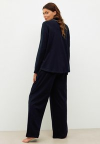 Woman with long wavy hair barefoot, wearing loose-fitting dark navy long-sleeve top and wide-leg pants, standing against a plain background.