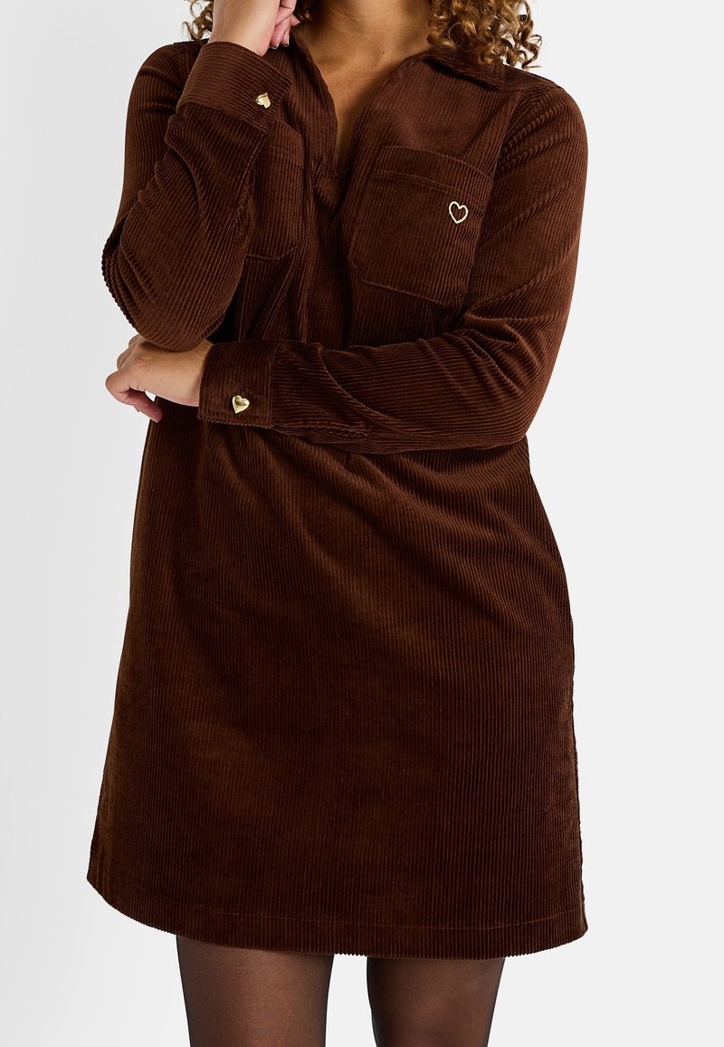 Femme portant une robe en velours côtelé marron avec des boutons dorés en forme de cœur et une broderie en forme de cœur sur la poche poitrine, posant avec un bras croisé.