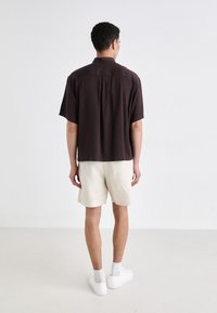 Camicia marrone scuro a maniche corte, vestibilità ampia, con pieghe sul retro. Abbinata a pantaloni corti beige chiaro e sneakers bianche. Design minimalista.