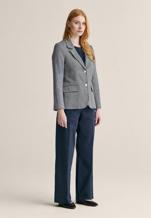 Rødhåret kvinde står iført en grå blazer, marineblå vide bukser, sorte sko og en mørk top mod en ensfarvet beige baggrund.