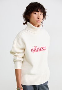 Cremefarvet sweater med høj, ribbet turtleneck. Har rødt broderet "ellesse" logo og ribbede detaljer på ærmer og kant.