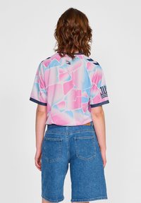 Osoba z falującymi włosami do ramion, ubrana w różowo-niebieską koszulę z krótkim rękawem w wzory oraz wysokie jeansowe szorty, zwrócona tyłem.