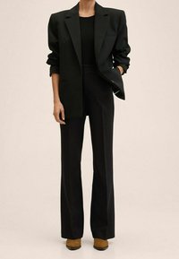 Zwarte blazer met opgerolde mouwen, gecombineerd met een aansluitend zwart topje en wijde zwarte broek. Tan schoenen, minimale hardwaredetails.