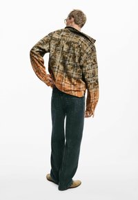 Chemise en flanelle à carreaux marron et noir avec un dégradé orange, poignets à boutons, associée à un jean large vert foncé et des chaussures beiges.