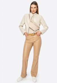 Una donna in piedi con una giacca di pelle beige, pantaloni in pelle ecologica color cammello, sneakers bianche e una borsa a tracolla beige.