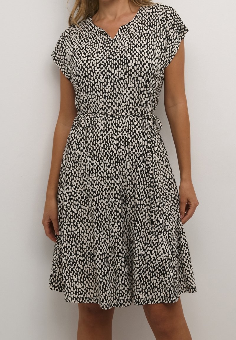 Mustavalkoinen kuviollinen mekko, jossa on V-pääntie, lyhyet hihat, naruhihnalla säädettävä vyötärö ja levenevä helma. Sileä kangas, jossa on pisteitä.