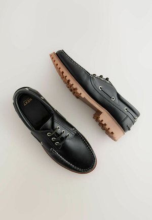 Fekete bőrcipők bézs gumi talppal és fűzőkkel, varrott díszítéssel és klasszikus csónakcipő dizájnnal.