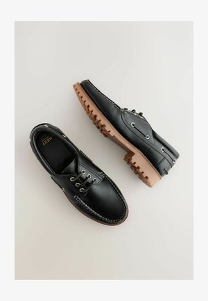 Fekete bőrcipők bézs gumi talppal és fűzőkkel, varrott díszítéssel és klasszikus csónakcipő dizájnnal.