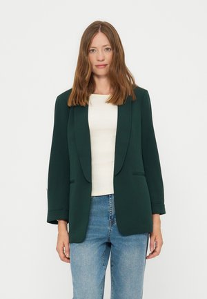 Vrouw met schouderlang bruin haar die een donkergroene blazer draagt over een witte top en blauwe spijkerbroek, staand tegen een effen lichte achtergrond.