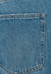 Tela de mezclilla de un tono azul claro, con un único bolsillo trasero delineado con costura naranja en contraste, textura plana y acabado limpio.