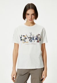 T-shirt en coton blanc arborant un graphique des personnages emblématiques de Warner Bros., avec le texte "100 ans de Warner Bros." sur le devant.