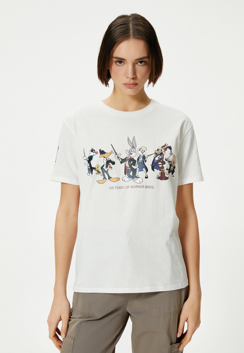 T-shirt en coton blanc arborant un graphique des personnages emblématiques de Warner Bros., avec le texte "100 ans de Warner Bros." sur le devant.