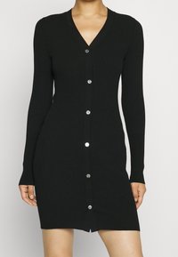 Robe cardigan en maille côtelée noire avec un col en V, des manches longues et des boutons argentés sur le devant, avec un ourlet qui tombe au-dessus du genou.