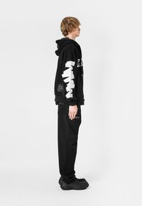 Sudadera negra con gráfico blanco en las mangas, combinada con pantalones negros y zapatos negros texturizados. Características incluyen bolsillos laterales y un corte relajado.