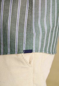 Grün gestreiftes Hemd mit blauen und weißen Linien, versehen mit einem dunkelblauen Etikett am Saum, kombiniert mit hellbeigen Cordhosen. Strukturierter Stoff sichtbar.