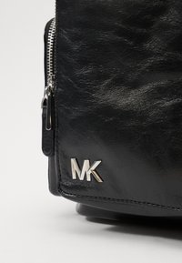 Bolso de cuero negro texturizado con detalle del logo "MK" en plata y cremallera. Presenta un diseño elegante y un acabado suave.