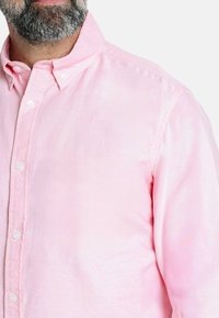 Camisa de botones de color rosa claro con una tela suave, que presenta un cuello clásico, costuras visibles y botones blancos estándar a lo largo del frente.