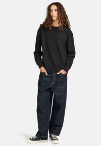Felpa nera con colletto rotondo e polsini a costine, abbinata a jeans in denim blu scuro larghi e scarpe da ginnastica nere con suole bianche.