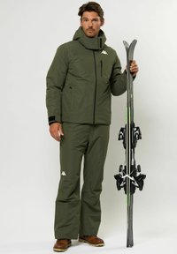 Olíva zöld síruha, pufferekből készült kabáttal és hozzá illő nadrággal. Figyelemre méltó részletei közé tartozik egy cipzáras zseb és logóval díszített elemek. Sílécek a kézben.