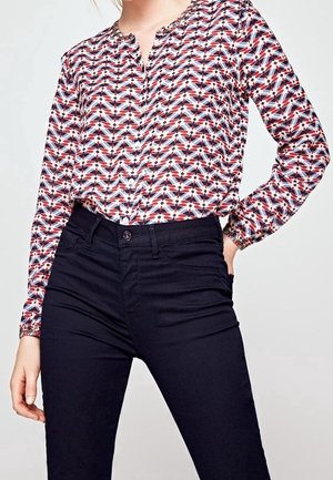 Žena v dlouhém rukávu vzorované blůze zastrčené do černých kalhot s vysokým pasem, ruka opřená o bok na jednobarevném pozadí.